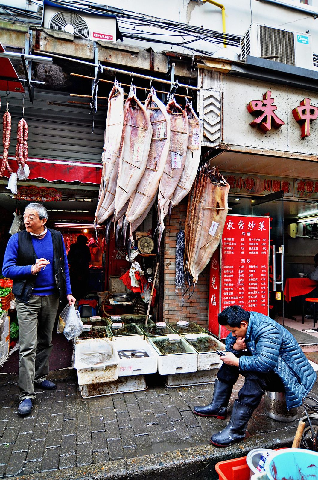 A Shanghai street market: split dried fish hang above tanks of live seafood, a red menu board on the wall, a man crouched checking his phone, another walking past with a bag.