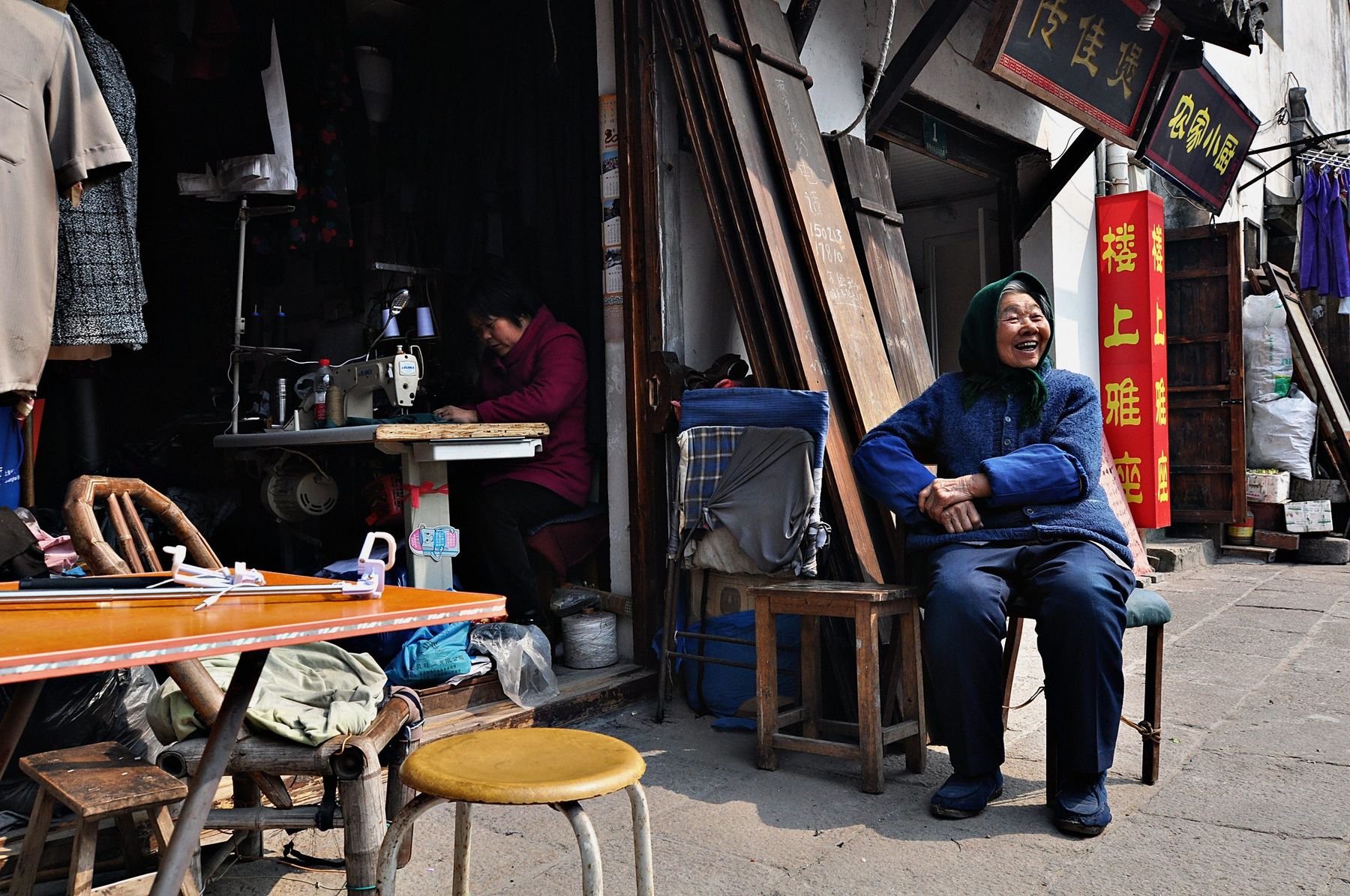 An old lane in Xinchang: a woman sews at a machine in a doorway while an older woman in a green headscarf sits outside laughing, red and gold shop signs above.