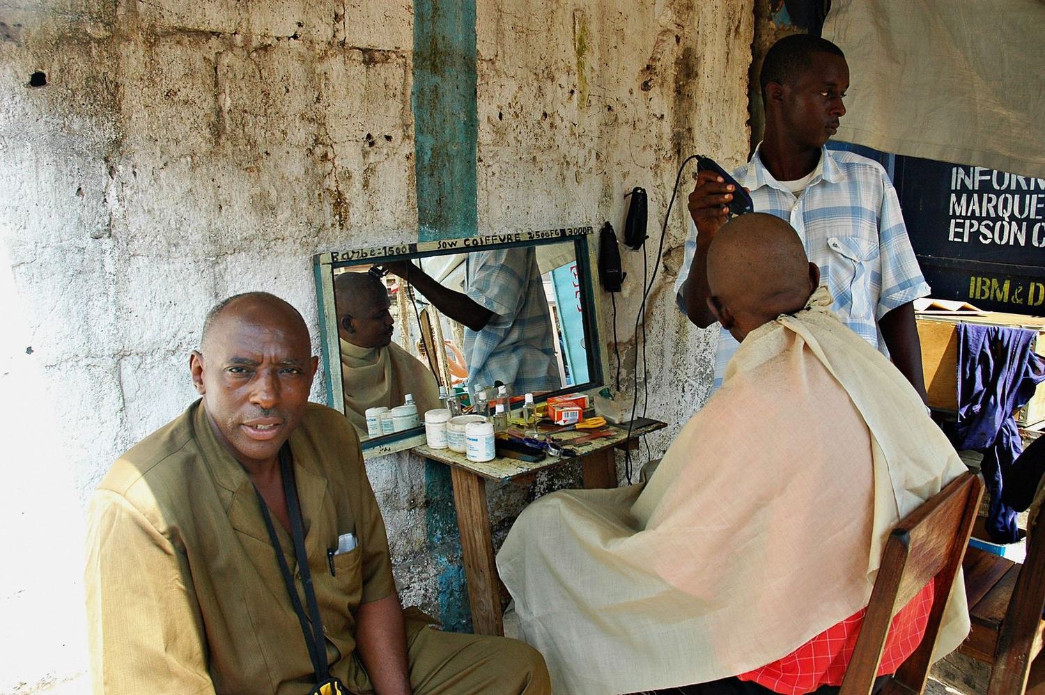 An open-air barbershop: one man waits in the foreground while a barber clippers another man&rsquo;s head, a hand-painted mirror and price list on the wall behind.