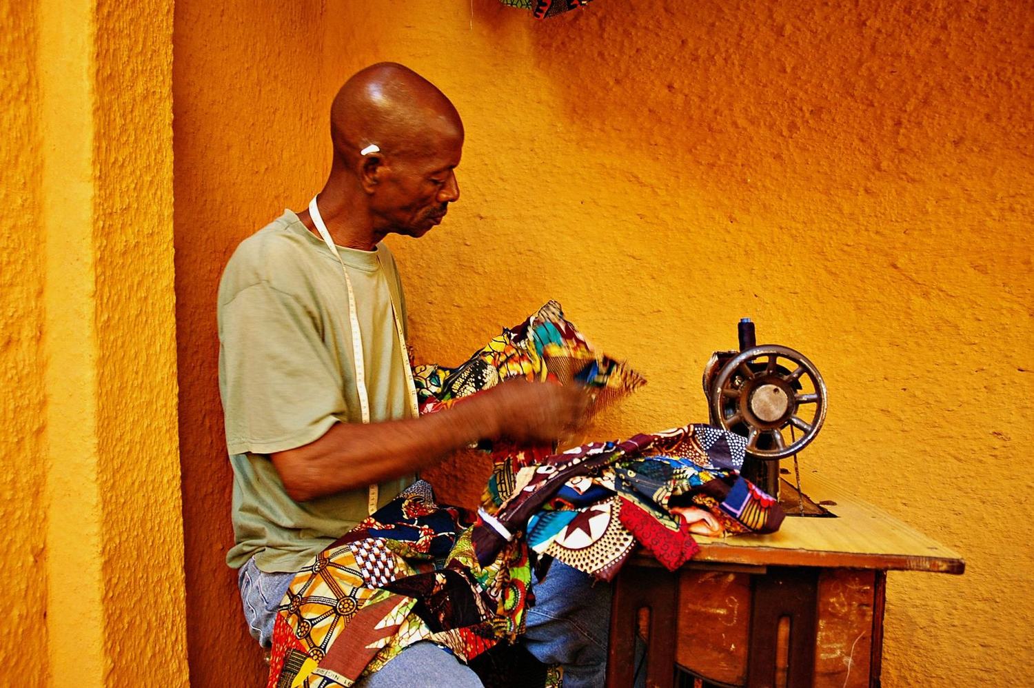 A tailor in a green t-shirt with a tape measure round his neck feeds bright wax-print fabric through a hand-cranked sewing machine against an ochre wall.