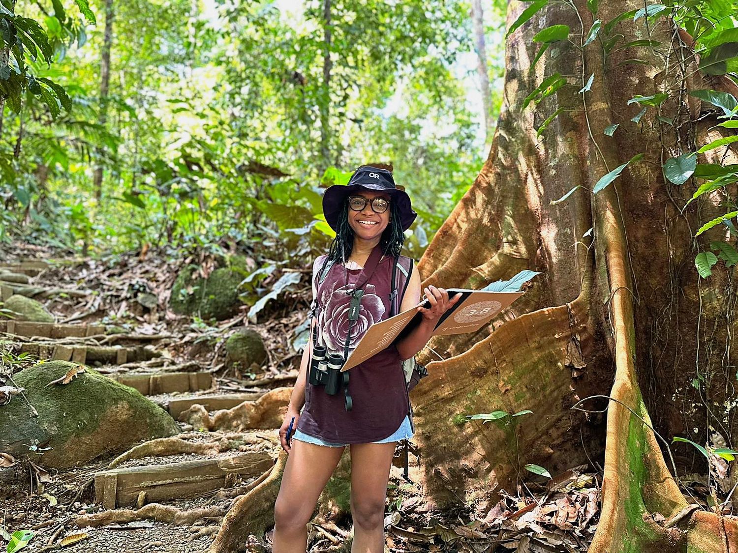 Genell Zuciya smiling on a rainforest trail beside a huge buttressed tree, binoculars around her neck, a field folder in hand.