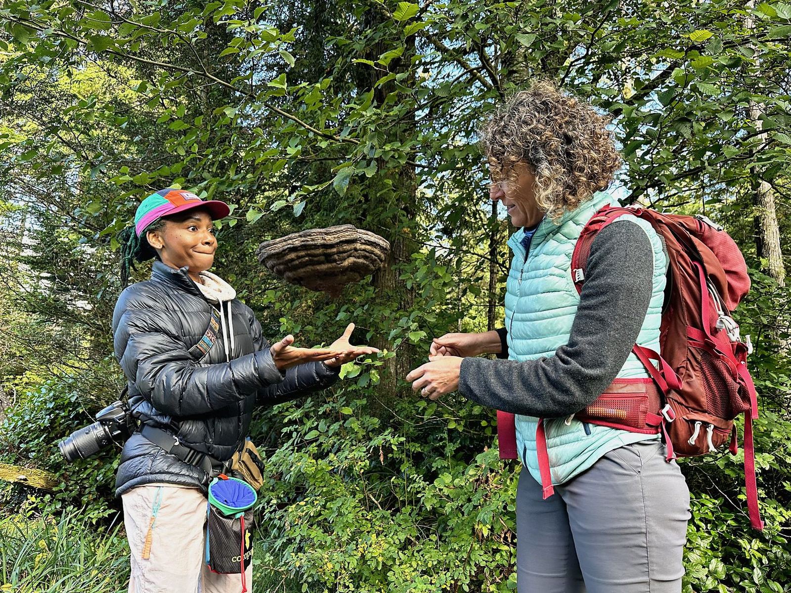 Genell tosses a large layered hoof fungus into the air toward a laughing companion in a green forest.